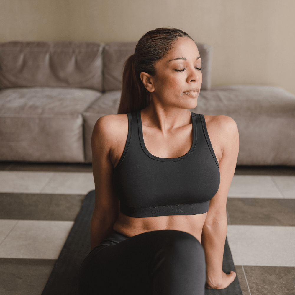 Woman wearing a black Gympak sports bra sitting on a yoga mat during a workout at home